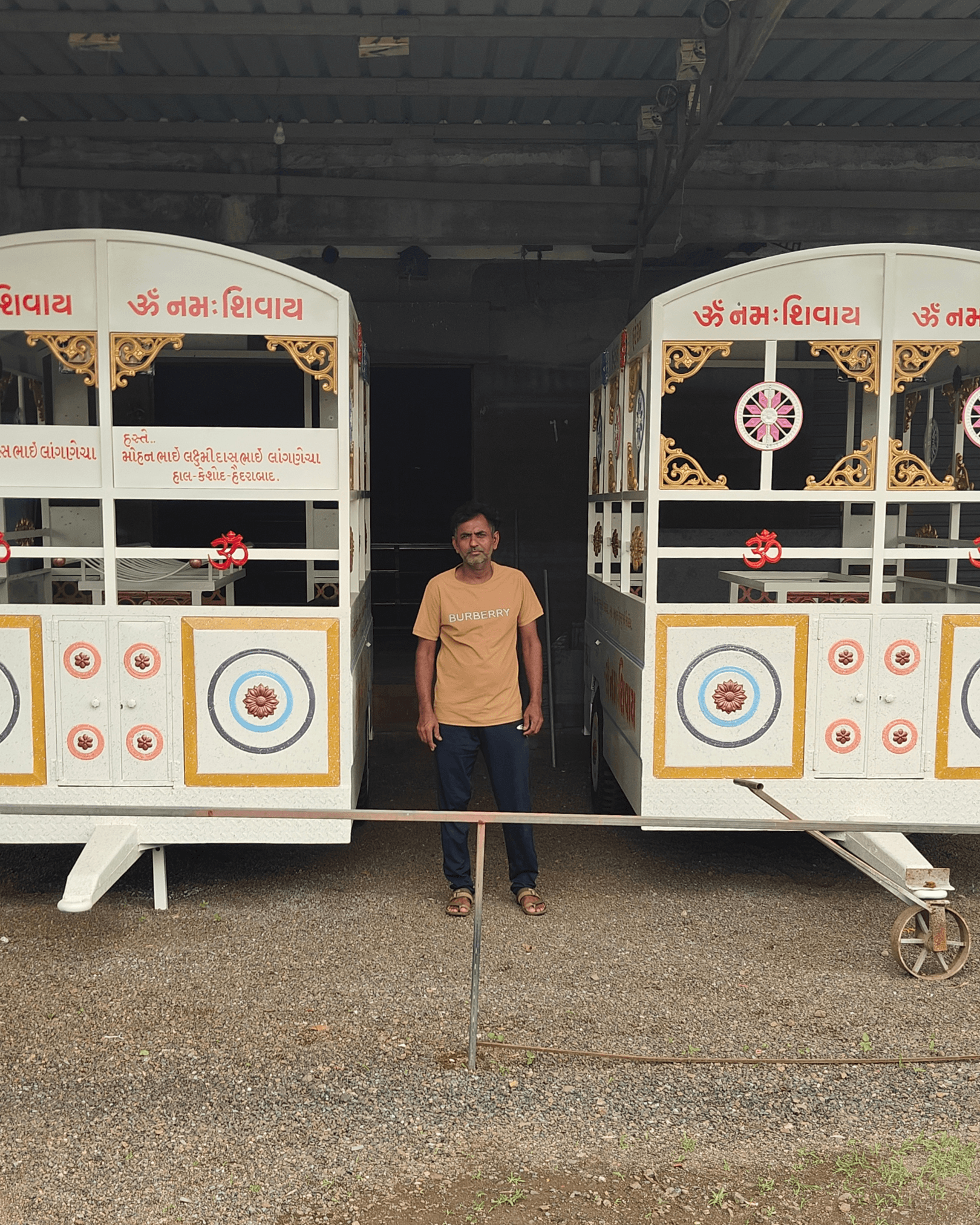 Founder of Jay Khodiyar Welding Works standing between two handcrafted Shanti Raths in the workshop